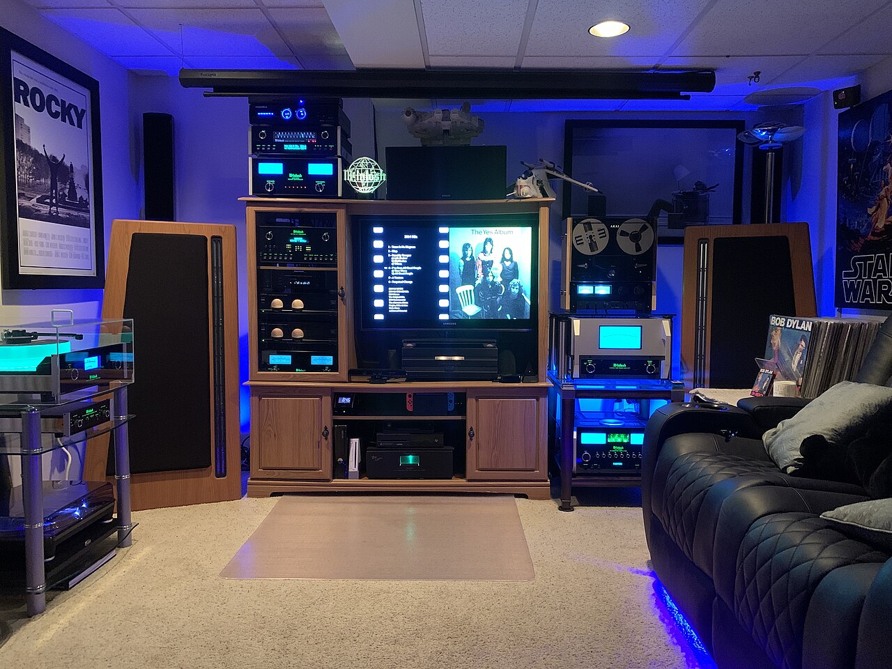 An audio equipment rack with amplifier level meters, floor-standing speakers either side of a large display, and seating